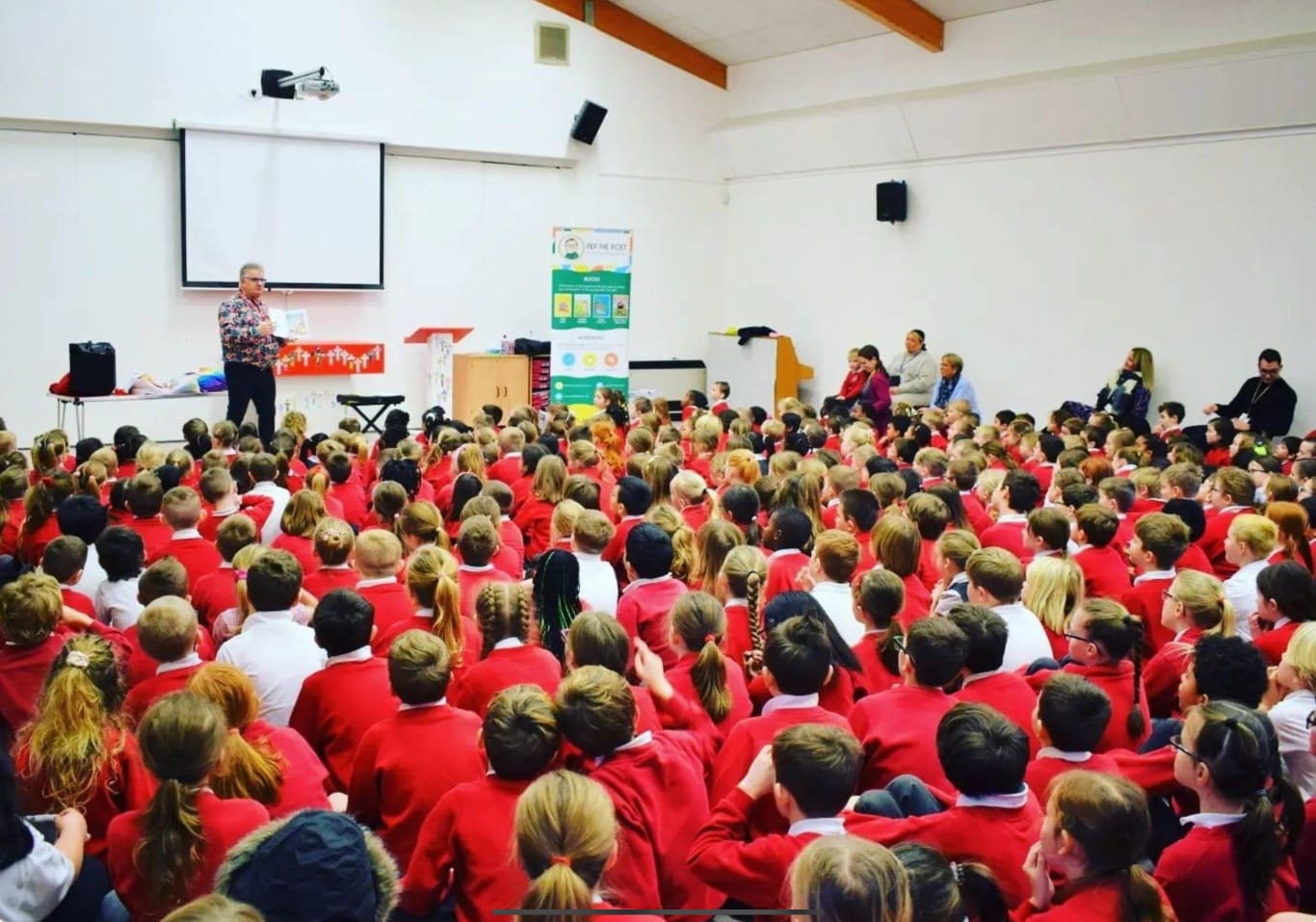 Pep the Poet standing at the front of an assembly hall full of children in red uniforms